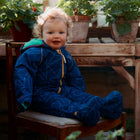 A baby wearing a navy EcoSplash Waterproof Fleece Lined Puddle Suit, seated on a chair in a greenhouse surrounded by potted plants.